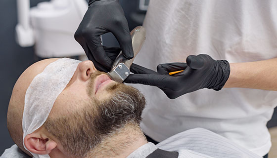 A man receives a beard trim from a barber in a well-lit barbershop.