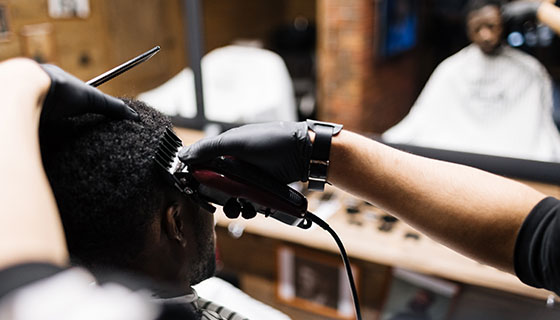 A man sits in a barber's chair while a barber trims his hair with scissors.
