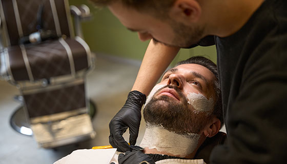 A man sits in a barber's chair as a barber carefully shaves his beard with a straight razor.