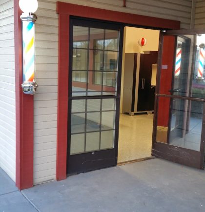 Entrance to a barber shop featuring glass doors, a classic striped barber pole, and an illuminated sign above.