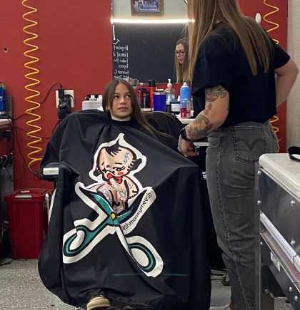 A hairstylist wearing a black shirt stands beside a client in a salon, with colorful hair products and a playful cape featuring a cartoon design.