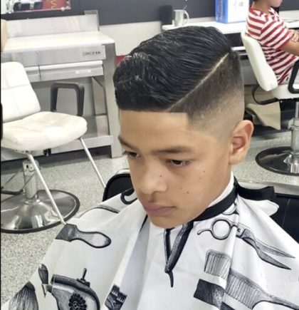 A young boy with a fresh barber's haircut sits in a salon chair, smiling at his reflection in the mirror.